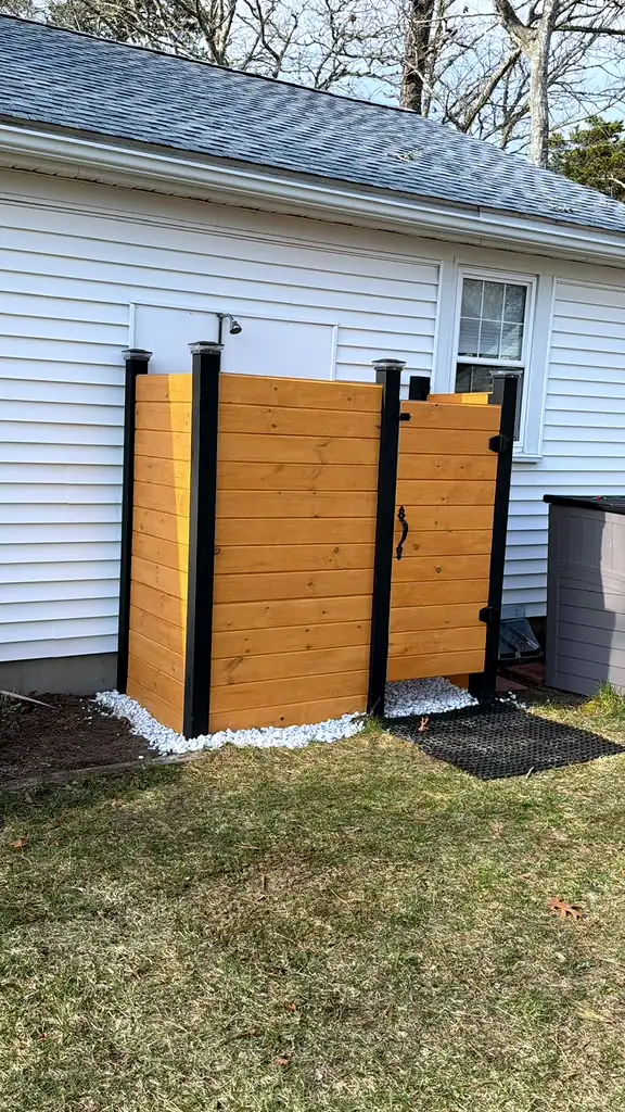 New horizontal wood outdoor shower enclosure with black posts beside a white house